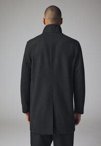 Zwarte wollen jas met een hoge kraag en lange mouwen, met een rechte snit, strakke lijnen en geen zichtbare knopen of versieringen.