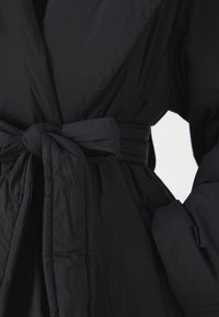 Abrigo tipo puffer negro con cintura ceñida, tela texturizada, mangas anchas y bolsillos laterales con acabado liso.