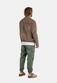 Chaqueta de ante marrón con cuello, combinada con pantalones cargo verdes con bolsillos laterales y zapatillas marrones con rayas blancas.