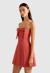 Frau trägt ein trägerloses, korallenrosa Minikleid mit großem Schleifendetail auf der Brust und steht vor einem schlichten, hellen Hintergrund.
