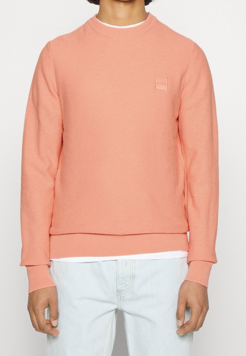 Personne portant un pull léger corail à manches longues et col rond par-dessus une chemise blanche avec un jean bleu clair, debout devant un fond uni.