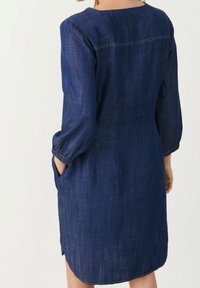 Vestido de mezclilla en azul oscuro, hasta la rodilla, con mangas tres cuartos, puños fruncidos, bolsillos laterales y un sutil detalle de costura en la parte posterior.