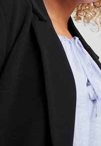 Czarna, fakturowana marynarka z prążkowanym wzorem, częściowo na jasnoniebieskiej koszuli z okrągłym dekoltem i ozdobą w postaci sznurka.