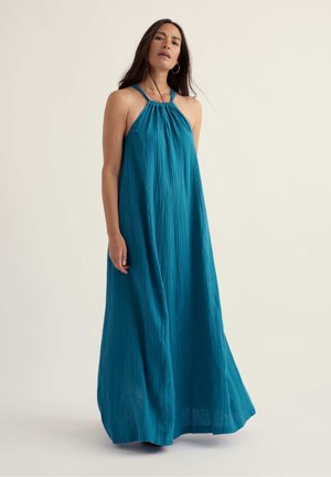 Donna che indossa un vestito lungo senza maniche color verde acqua con collo halter, in piedi contro uno sfondo semplice.