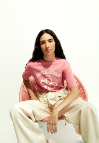 Roze katoenen t-shirt met het "ARMEDANGELS" logo en een bloemenafbeelding. Model draagt een crèmekleurige, wijde broek en zit in een ronde roze stoel tegen een witte achtergrond.
