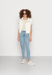 Colete cremoso acolchoado sobre uma t-shirt listrada em verde e branco, combinado com jeans rasgados azul claro e ténis bege. Óculos de sol redondos pretos incluídos.