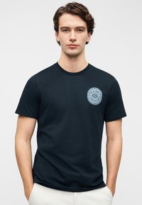 Μπλούζα από βαμβάκι σε ναυτικό μπλε χρώμα με κοντά μανίκια και στρογγυλή λαιμόκοψη, με κυκλικό ανοιχτό μπλε λογότυπο στην αριστερή περιοχή του στήθους. Ομαλή υφή.