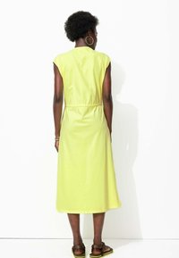 Robe jaune vif, longueur genoux, à manches courtes. Elle présente une taille élastique, une coupe décontractée et un léger éclat. Portée avec des sandales marron.