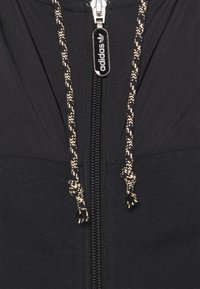 Svart jacka med dragkedja och kontrasterande texturerad huva samt snoddar, med en Adidas-logga på dragkedjan.