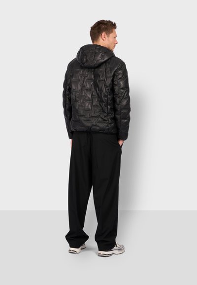 Homme portant une veste à capuche noire matelassée, un pantalon noir ample et des baskets blanches, se tenant avec les mains dans les poches, tourné le dos.
