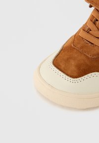 Καφέ σουέντ αθλητικό παπούτσι με μύτη από  κρεμ καουτσούκ, ραμμένα στοιχεία και υφή στη σόλα. Διαθέτει παραδοσιακό κλείσιμο με κορδόνια και επενδυμένο κολάρο.