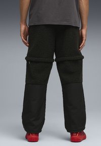 Pantaloni neri foderati in pile con materiale testurizzato e soffice e pannelli inferiori a contrasto in tessuto liscio. Scarpe da ginnastica rosse visibili.