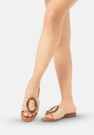 Piernas femeninas que llevan sandalias beige de tejido y estilo deslizante con una hebilla ovalada grande de madera, de pie sobre una superficie gris clara.