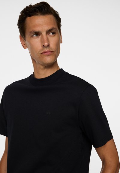 Uomo con capelli ricci corti che indossa una semplice maglietta nera a girocollo, che guarda leggermente a sinistra su uno sfondo bianco.