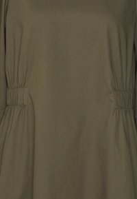 Túnica verde oliva de corte relajado, con ruches laterales, mangas largas y una suave mezcla de algodón. Diseño minimalista.