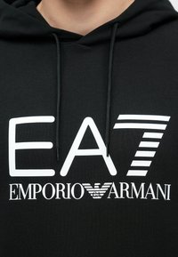 Fekete kapucnis pulóver pamutból. Nagy fehér nyomtatással, rajta az "EA7" és "EMPORIO ARMANI" feliratokkal. A húzózsinórok fekete sz�ínűek, texturált anyagból készültek.