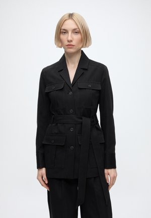 Kvinde med glat blond bobfrisure iført en sort jakke med bælte og lommer med knapklap, stående mod en hvid baggrund.