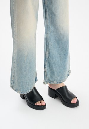 Mules en cuir noir à bout ouvert avec un talon épais et une texture lisse, assorties à un jean large bleu clair déchiré.