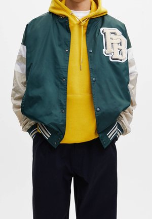 Personne portant une veste varsity verte et beige par-dessus un sweat à capuche jaune avec un pantalon foncé, debout les mains dans les poches.