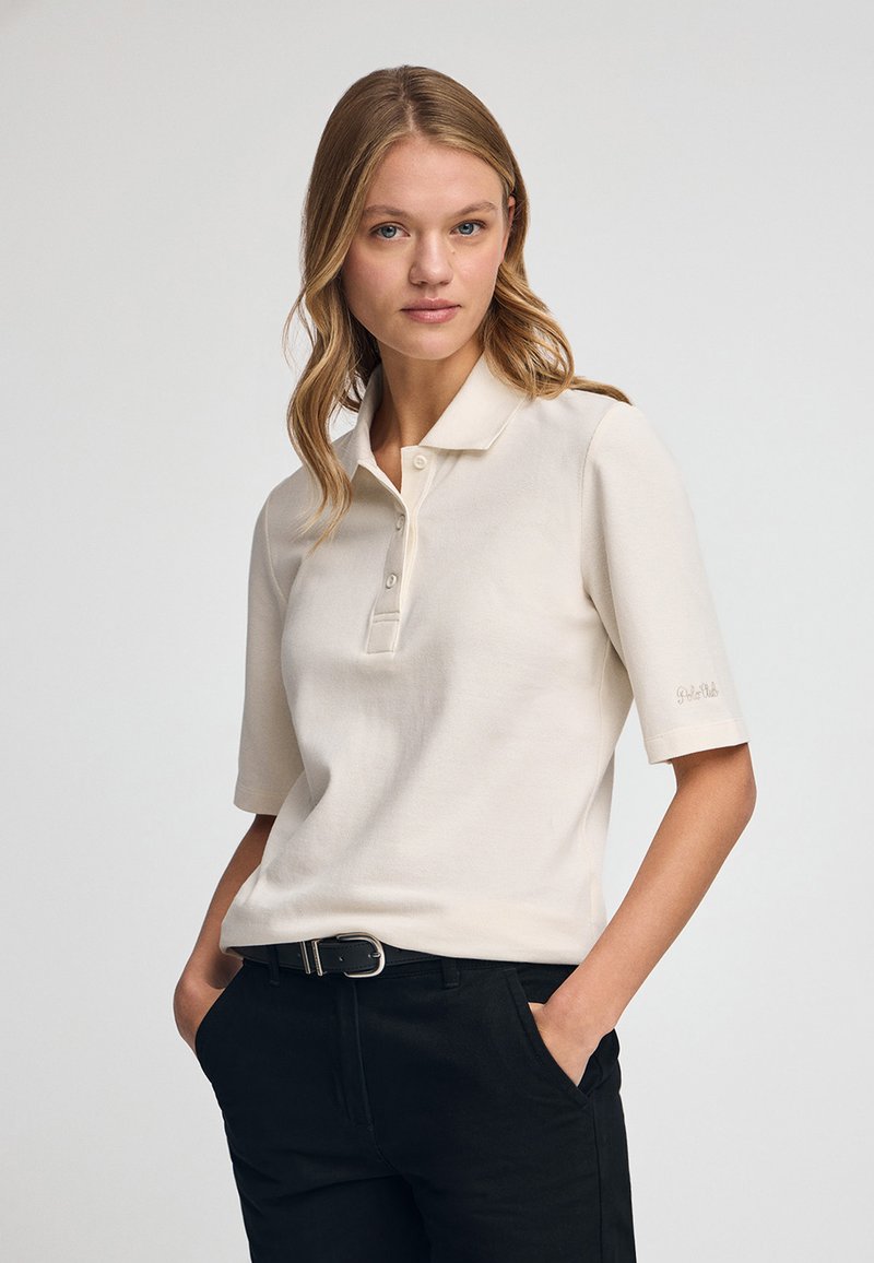 Korte mouw polo shirt in licht beige, gemaakt van zachte stof. Heeft een kraag, een knoopsluiting met drie knopen en een geborduurd logo op de mouw.