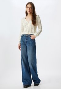 Cremefarbener grob gestrickter Cardigan mit tiefem V-Ausschnitt, kombiniert mit hoch taillierten, weit geschnittenen blauen Jeans. Sanfte Textur und entspannter Schnitt.