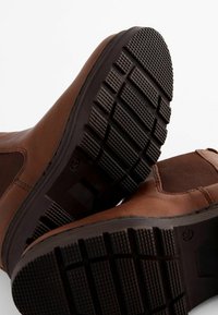Bottines en cuir marron avec semelles texturées en caoutchouc marron. Dotées de panneaux latéraux élastiques pour un enfilage facile. Design robuste et durable.