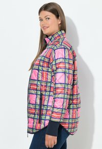 Kvinde iført en farverig ternet quiltet jakke med pink, orange, grønne og navy striber, der står sidelæns mod en hvid baggrund.