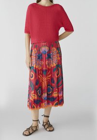 Un pull en tricot rouge court associé à une jupe plissée à fleurs arborant des motifs orange, rose et vert foncé. Sandales brunes à lanières.
