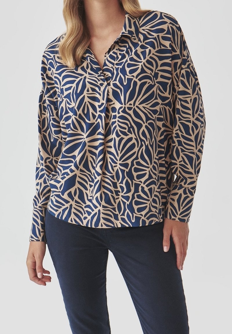 Femme portant une chemise à manches longues bleu et beige à boutons avec un motif abstrait de feuilles et un pantalon bleu foncé.