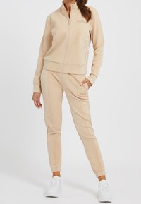 Chándal beige hecho de tela suave, con chaqueta con cremallera y pantalones ajustados con costuras de contraste y acentos de logo. Combinado con zapatillas blancas.
