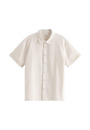 Teenagedreng stående med hænderne i lommerne iført en beige kortærmet skjorte, hvid t-shirt, marineblå bukser og sorte sandaler på en hvid baggrund.