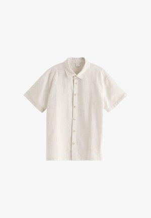 Teenager-Junge steht mit eingeschobenen Händen in den Taschen, trägt ein beiges kurzärmeliges Hemd, ein weißes T-Shirt, marineblaue Hosen und schwarze Sandalen vor weißem Hintergrund.