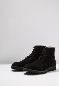 Botines de ante negro con cordones y cremallera lateral. Suela de goma resistente con suela antideslizante, textura superior suave y detalles de costura mínimos.
