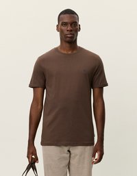 Brun bomulds t-shirt med rund halsudskæring og opfoldede ærmer. Har et lille blåt logo på brystet. Båret med lyse fløjl bukser.