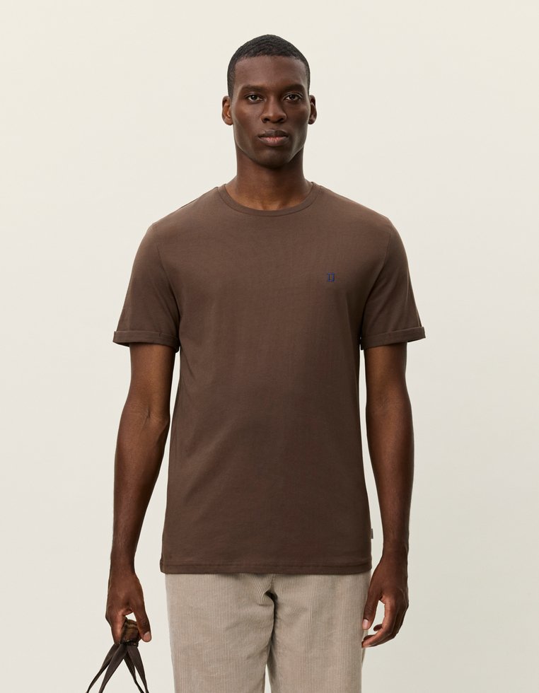 Brun bomulds t-shirt med rund halsudskæring og opfoldede ærmer. Har et lille blåt logo på brystet. Båret med lyse fløjl bukser.