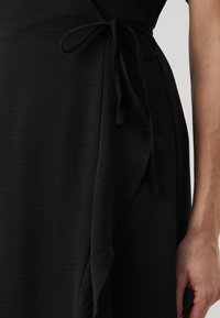 Black dress with a textured fabric, featuring a wrap design, side tie closure, and a flowing shape. Shoulder and sleeve detail visible.