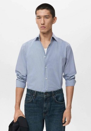 Camisa de botón en azul suave con rayas blancas, mangas largas remangadas, combinada con unos jeans de mezclilla oscuros y una chaqueta negra colgada de un brazo.