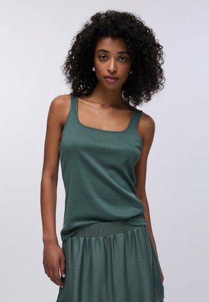 Donna con capelli ricci che indossa un top verde senza maniche e una gonna abbinata, in piedi davanti a uno sfondo grigio chiaro uniforme.