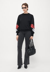 Person som har på seg en svart hettegenser med rød og hvit rose-design på ermene, mørkegrå jeans, svarte spisse støvler, solbriller, og holder en svart håndveske.