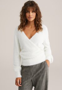 Valge, paksu villase sisse tõmmatud kampsun avara V-kaelusega ja pikkade varrukatega, koos tekstuursete hallide pükstega. Pehme, mugav kangas õrnalt särav.