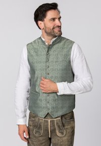Grün gemusterte Weste mit mandarinischem Kragen, silbernen Knöpfen und zwei Fronttaschen, kombiniert mit einem weißen Hemd und braunen Hosen.
