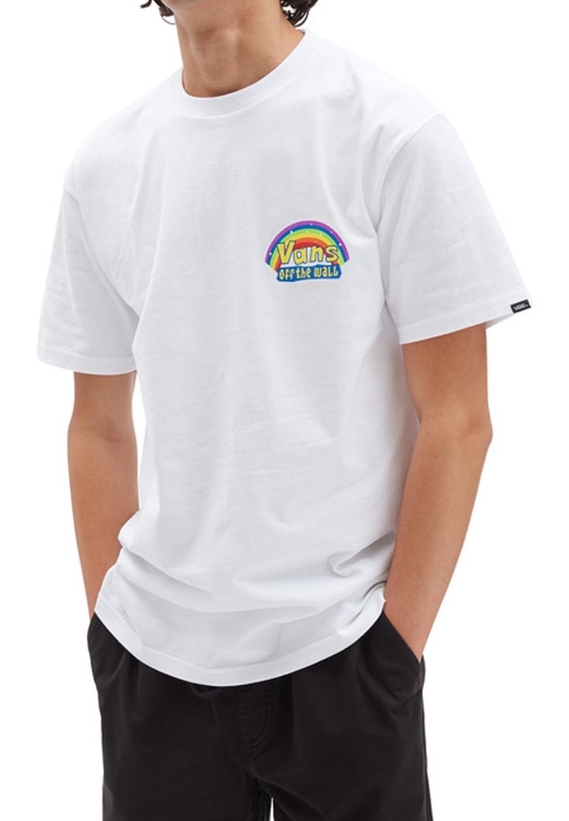 Eine Person trägt ein weißes Vans-T-Shirt mit einem kleinen Regenbogenlogo und dem Text "Vans Off the Wall" auf der Brust, die Hände in den Taschen von schwarzen Shorts.
