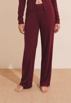 Person wearing loose maroon pajama pants and matching long-sleeve top standing barefoot on beige carpet.