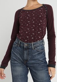 Maroon långärmad topp med blommönster, svart piping och knappdetaljer; matchad med högmidjade blå denimjeans.