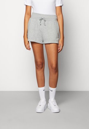 Kobieta w jasnoszarych szortach z troczkiem, białych skarpetkach ze ściągaczem z czarnymi logo oraz białych tenisówkach, stojąca na tle jednolitego tła.