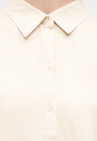Κοντινή λήψη ενός πουκάμισου από λινό εκρού χρώματος με μυτερό γιακά και τέσσερα ορατά λευκά κουμπιά στο μπροστινό μέρος.