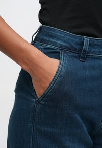 Mão de tom de pele escura colocada dentro do bolso frontal de uns jeans de ganga azul escuro, usados com uma blusa preta.