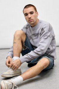 Unge mand sidder på gulvet og binder snørebåndene, iført en grå sweater med kæde-mønster, denim shorts, hvide sokker og sneakers.