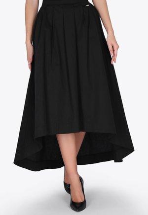 Jupe midi noire à taille haute et ourlet descendant avec des plis, portée par une personne associée à des escarpins noirs à bouts pointus, les mains détendues le long du corps.