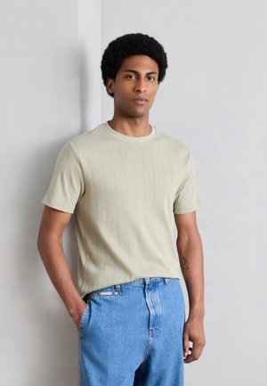 Mladý muž s afro účesom, ktorý má na sebe béžovú textúrovanú košeľu s krátkym rukávom a modré džínsy, stojí pri svetlo sivej stene.